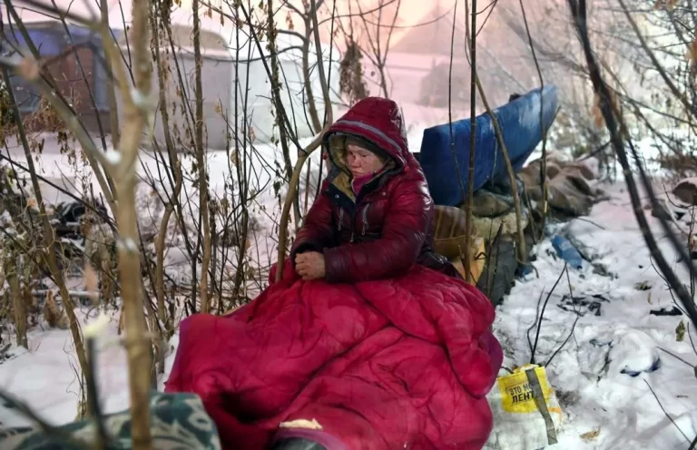 A person sitting on the ground outdoors in winter, wrapped in a red blanket, surrounded by bare trees and snow.