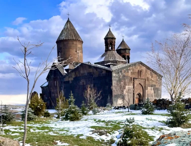 A historic stone church with three pointed towers stands on a partially snow-covered landscape with some greenery and barren trees, under a cloudy sky.