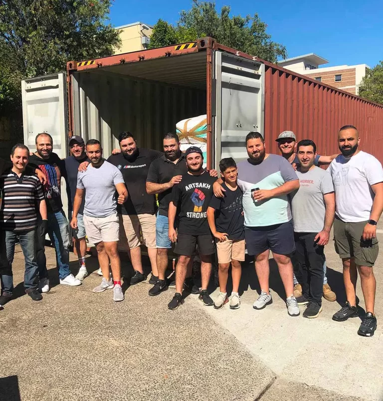 A group of thirteen men stands in front of an open shipping container outdoors on a sunny day, smiling and posing for the photo. A few have their arms around each other's shoulders.