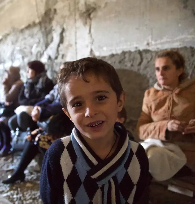 A young child in a patterned sweater stands in front of adults seated against a rough, concrete wall. The child is smiling, and the environment appears to be indoor and makeshift.