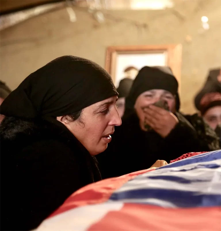 Women in black headscarves mourn beside a draped casket.