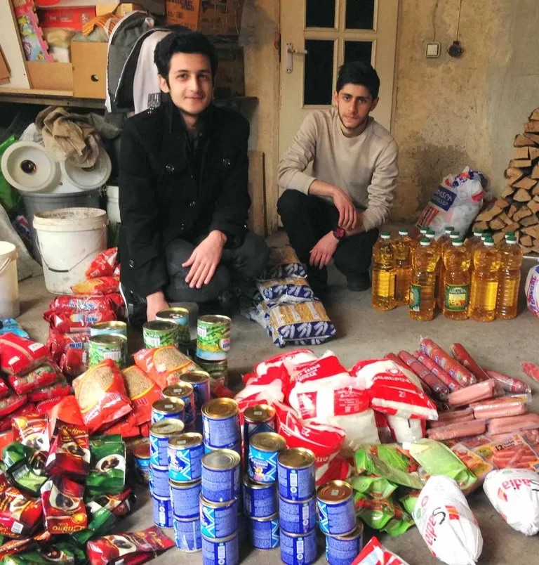 Two young men sit on a floor surrounded by a large assortment of groceries, including cans, bags of pasta, bottles of oil, and various other food items.