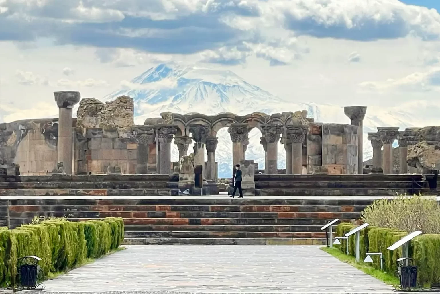 Ruins of an ancient structure with columns, set against a mountainous backdrop. People walk along a pathway leading to the site, surrounded by neatly trimmed greenery under a cloudy sky.