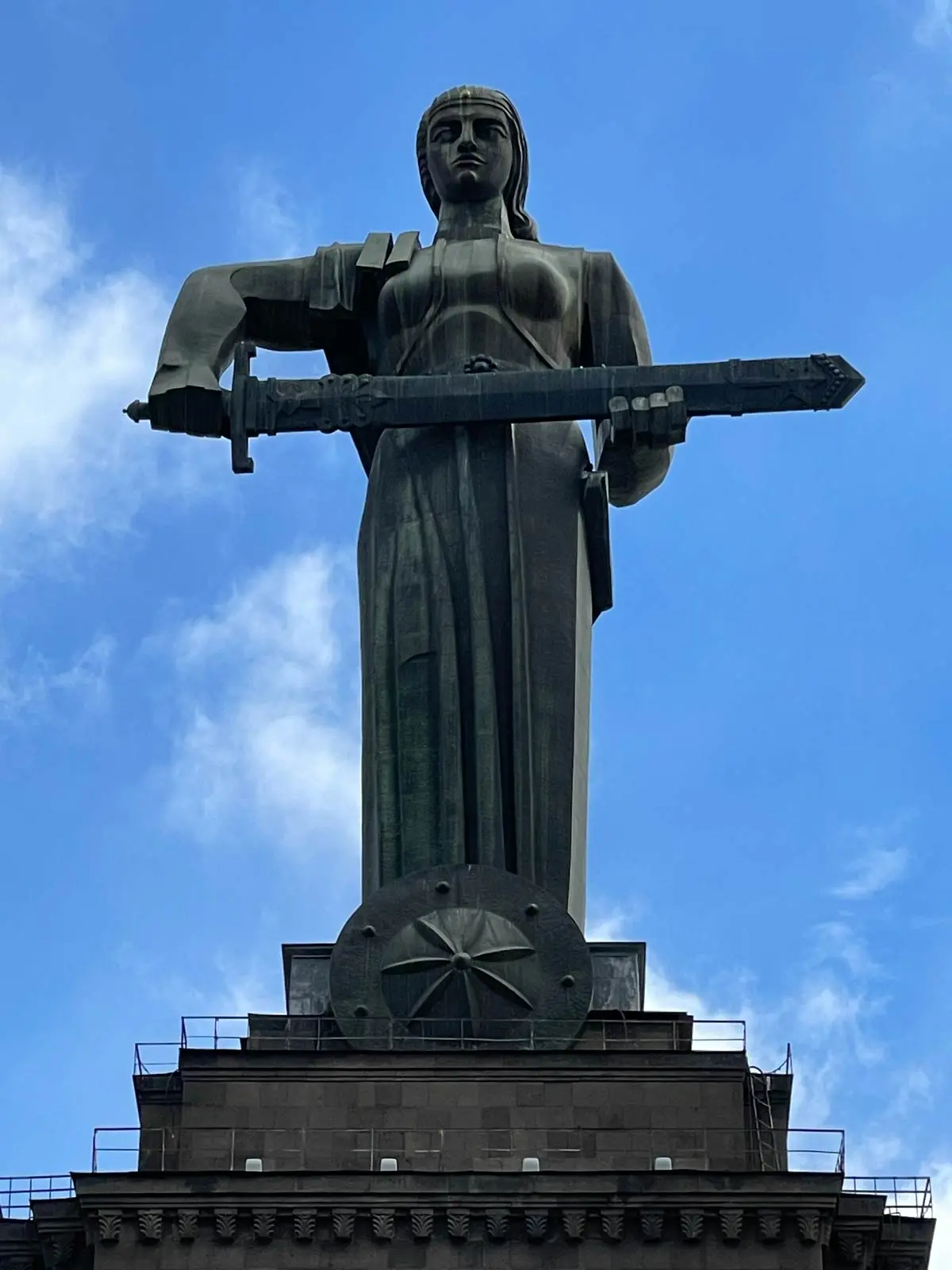 A large stone statue of a woman holding a sword stands atop a tall building against a blue sky with clouds.