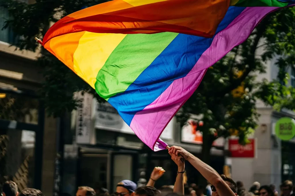 A large rainbow flag is being waved by individuals in an outdoor setting, surrounded by trees and buildings.