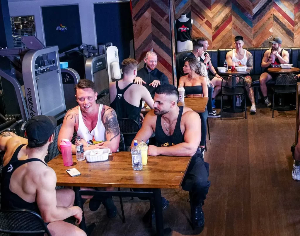 A group of people, mostly in workout attire, are seated around tables in a gym area with fitness equipment in the background. They appear to be conversing and enjoying drinks.