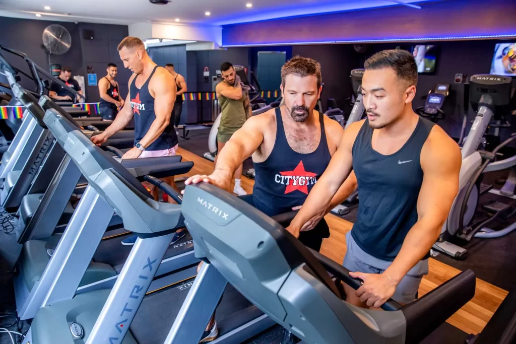 A group of men working out in a gym, some using treadmills and others performing various exercises. Two men in the foreground are focused on treadmill settings.