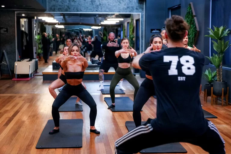 A fitness instructor leads a focused group of individuals in a squatting exercise at a gym, with mirrors lining one wall and potted plants decorating the room.
