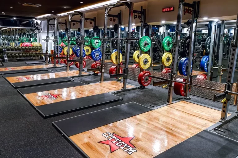 A well-lit gym features multiple weightlifting platforms with colorful weight plates, barbell racks, and mirrors. A digital clock shows 05:28. The floor is a mix of rubber and wooden surfaces.