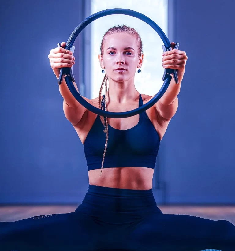 A woman in athletic wear sits in a split and holds a fitness ring in front of her, performing an exercise.