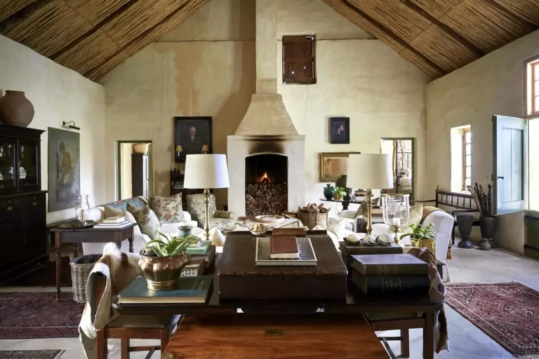 A rustic living room features a fireplace, wooden ceiling, various paintings, and cozy seating. The space is adorned with books, lamps, and decorative items, creating a warm and inviting atmosphere.