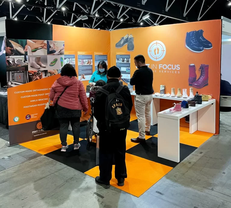 A group of people standing at an orange-themed booth titled "Feet in Focus Clinic Services" at a trade show, examining orthopedic shoes and related products displayed on tables.