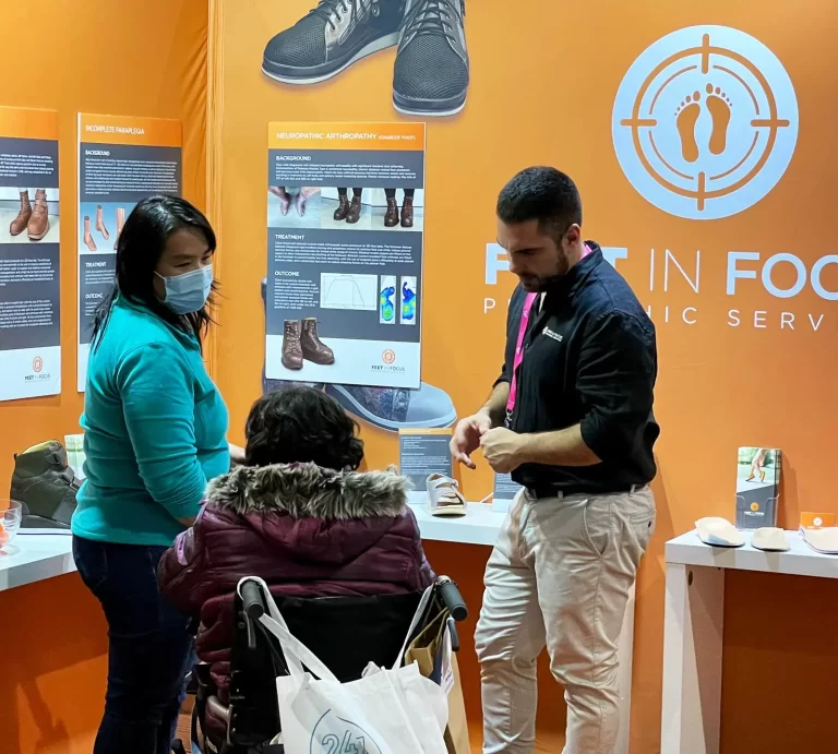 A man demonstrates foot care products to a woman in a wheelchair and another standing woman at a booth with an orange background and informational posters.
