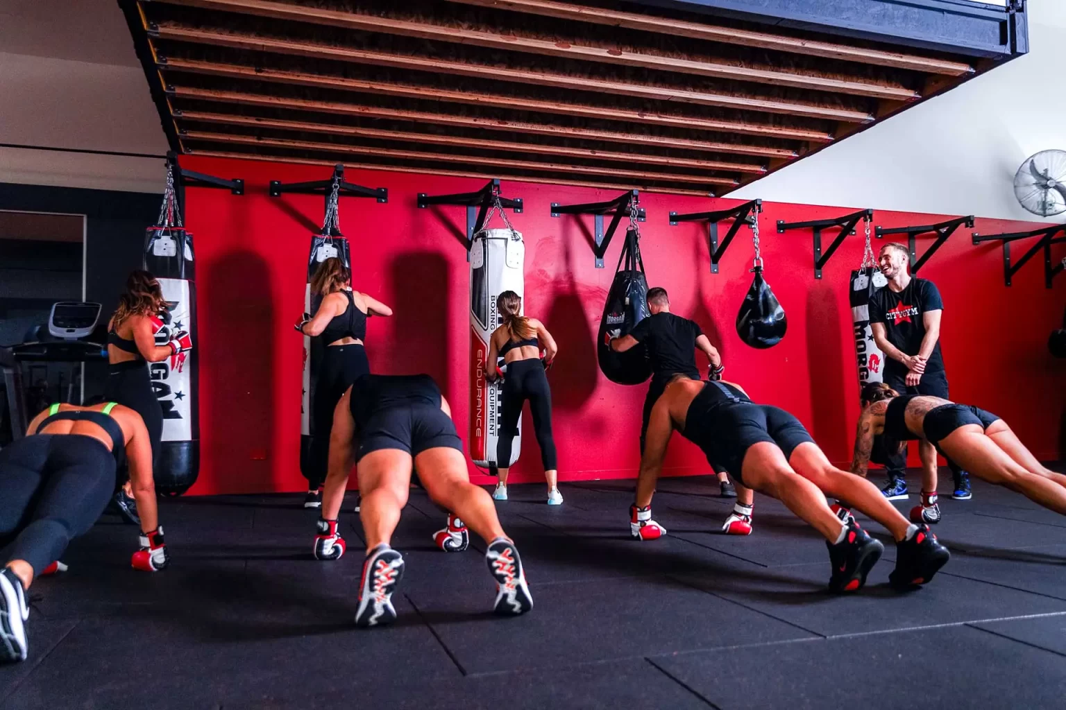 People in a gym performing various exercises, including push-ups and bag punching, with a trainer observing. Red wall with hanging bags in the background.