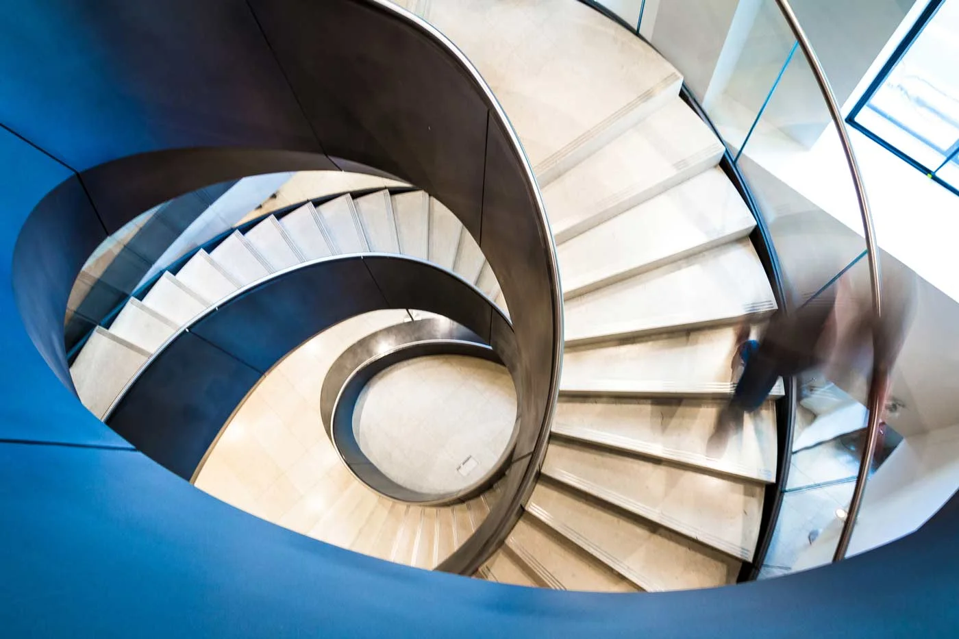 A person walks down a modern, spiral staircase with metallic railings and polished steps in a well-lit indoor setting.