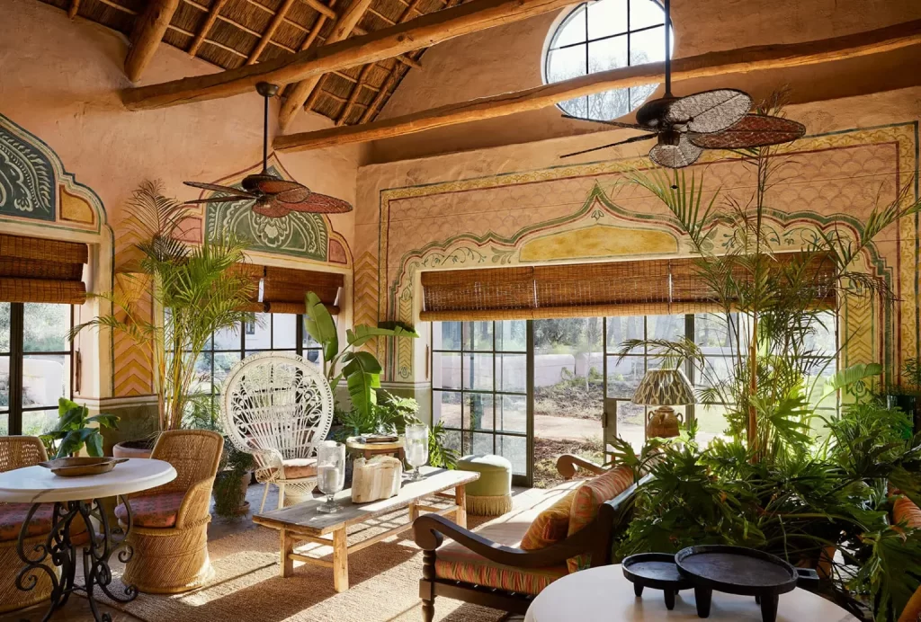 A sunlit room with a high, thatched ceiling, decorative wall art, large windows, wicker furniture, indoor plants, and ceiling fans.