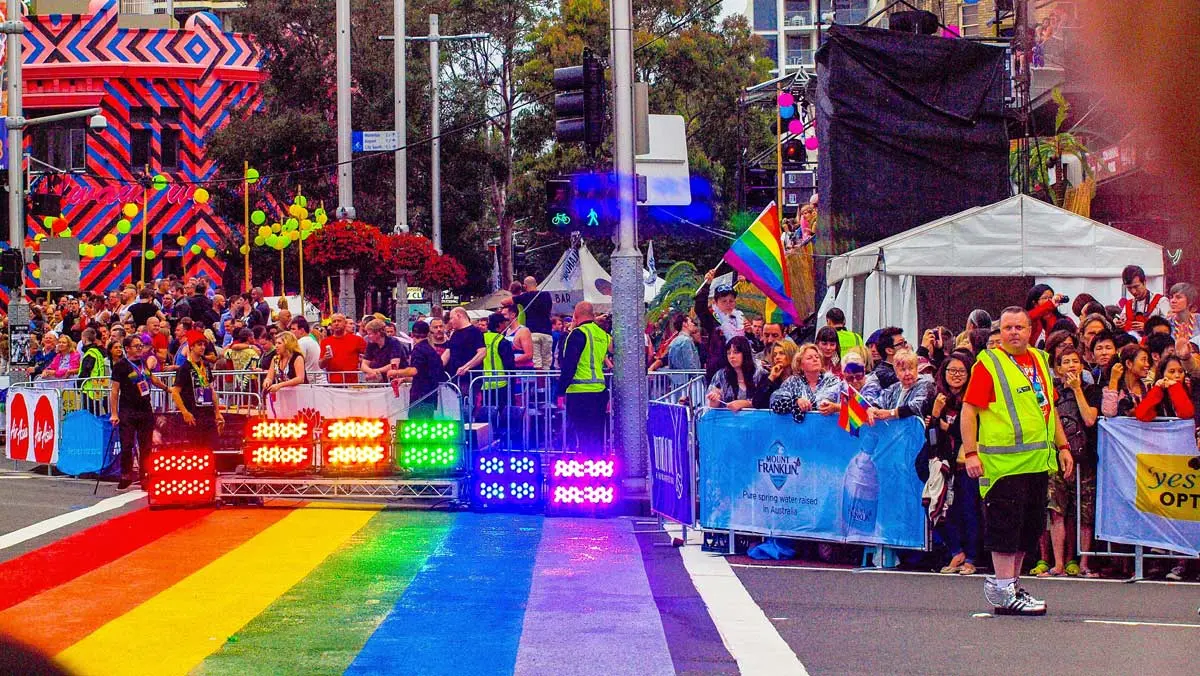 A bustling crowd at an outdoor event with a rainbow-striped street and colorful decor, supervised by event personnel in bright vests.