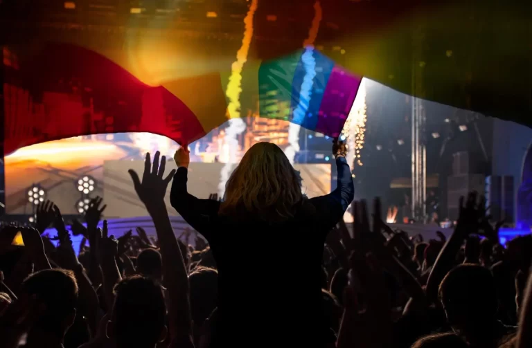 A person with raised hands is waving two rainbow flags at an indoor concert, surrounded by a cheering crowd and colorful stage lights.