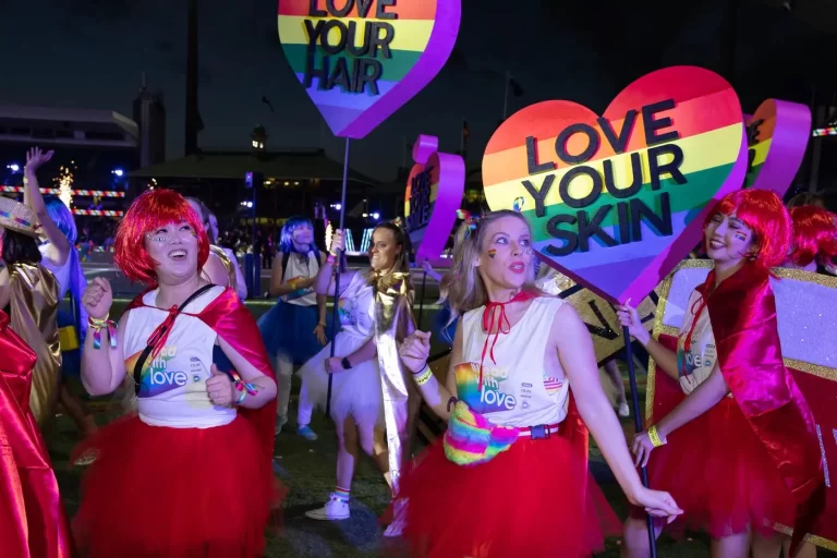 A group of people in matching outfits hold colorful heart-shaped signs that read "Love Your Hair" and "Love Your Skin" at a nighttime event.
