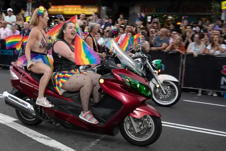 Participants riding motorcycles adorned with rainbow flags parade down a street, as an enthusiastic crowd watches and takes photos during a pride event.