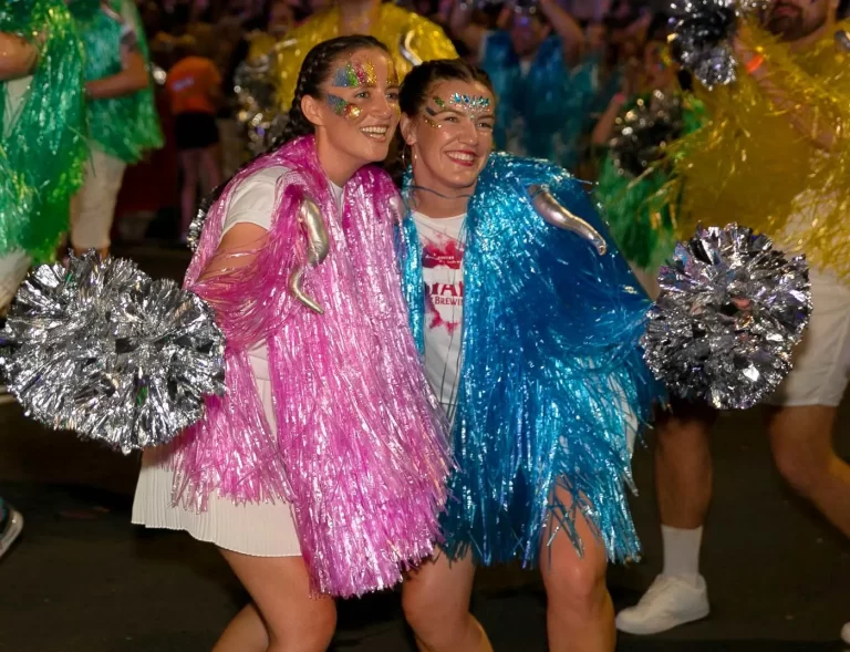 Two people dressed in colorful, sparkly outfits with pom-poms smile and pose for a photo during a festive event.