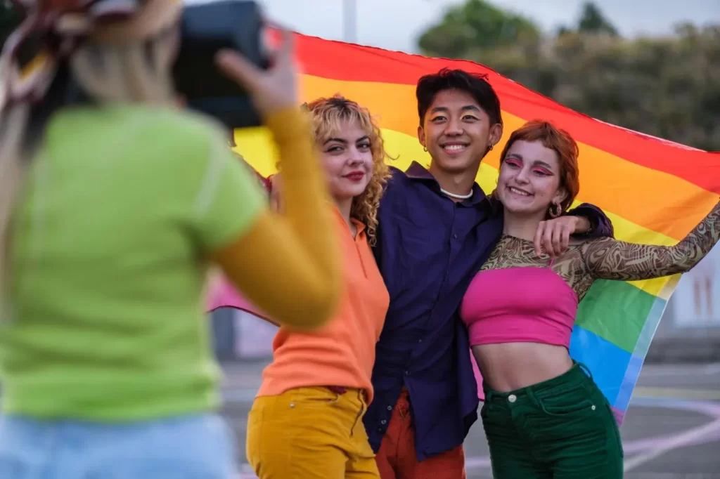 Three people pose for a photo while holding a rainbow flag. Another person takes their picture with a camera. They are outdoors in a casual setting.
