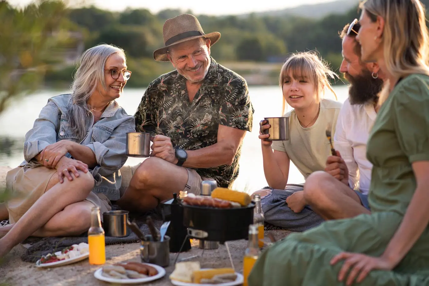 A group of people sit outdoors on a blanket near a portable grill, holding mugs and talking. Food items like corn and drinks are spread out in front of them.