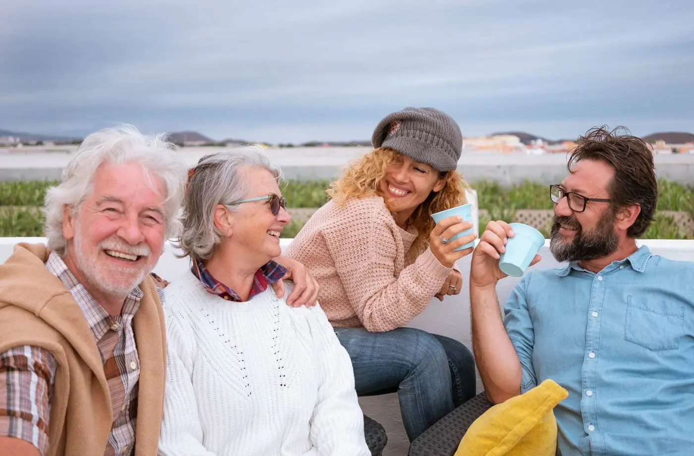 Four adults, two men and two women, sit together outdoors smiling and holding mugs. The background features an overcast sky and distant hills.