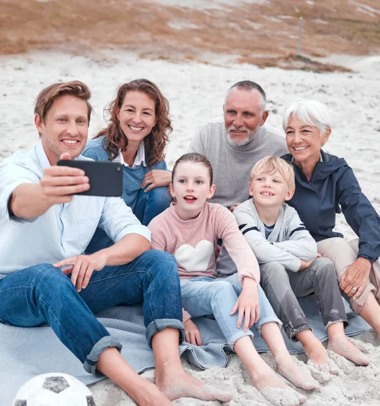 A group of six people, comprising multiple generations, sit together on a sandy beach, with one person taking a selfie. A soccer ball is visible in the foreground.